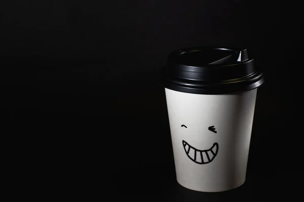 one white disposable coffee cup closed with a black lid with a smile on the body, stands on a black background, isolated on black.