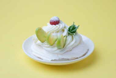 Beyaz tabakta Pavlova kremalı tatlı ve sarı arka plan. Kremalı, yuvarlak kremalı kremalı krema, nane yapraklı ve kirazlı tatlı.