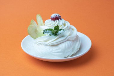 Beyaz tabakta Pavlova kremalı tatlı ve turuncu arka plan. Kremalı, yuvarlak kremalı kremalı krema, nane yapraklı ve kirazlı tatlı.