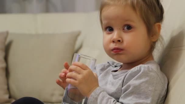 Little child drinking water from a glass sitting on a sofa in the room — Stock Video