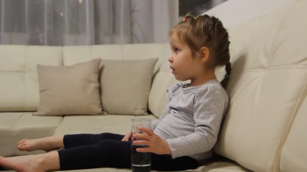 Niño bebiendo agua de un vaso sentado en un sofá de la habitación — Vídeos de Stock