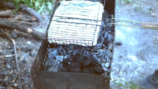 Un homme cuisine quelque chose dans du papier grillé tout en ajoutant des charbons dans un gril — Video