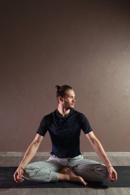 Genç sporcu yoga yapıyor, Yarım Lotus pozisyonunda meditasyon yapıyor spor kıyafetleri giyiyor, kapalı alanda uzun, gri yoga stüdyosu.