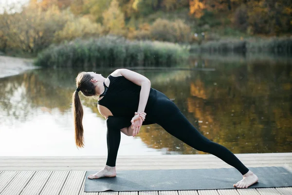 Mujer joven haciendo yoga asana en la naturaleza con vista al lago. Entrenamiento nocturno al aire libre, deportes y estilo de vida saludable — Foto de Stock