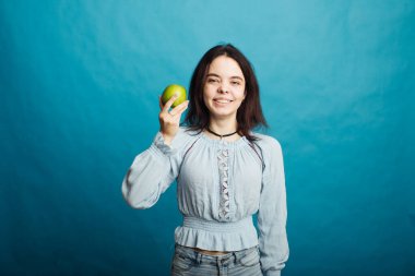 Mavi bir arka planda elinde yeşil bir elma tutan mutlu bir genç kız resmi.