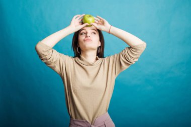 Mavi bir arka planda elinde yeşil bir elma tutan mutlu bir genç kız resmi.