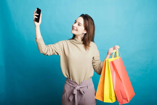 Mavi arka planda duran mutlu genç bir kız resmi. Alışveriş çantaları ve telefonla. — Stok fotoğraf