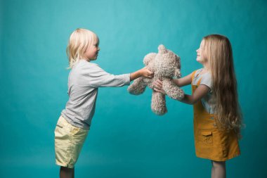 Mavi arka planda bir kız ve erkek, gri bir ayıyla el ele tutuşuyorlar. Çocuklar çok neşeli. Oyuncaklar