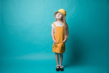 Mavi arka planda sarı şapkalı bir kız kameraya bakıyor. Kız sarı bir elbise giymiş.