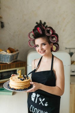 Önlüklü ve bigudili genç bir anne, küçük çocuğuyla birlikte mutfakta doğum günü pastası hazırlıyor.