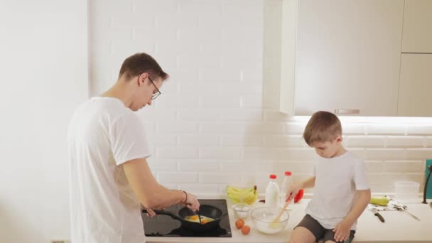Niño hijo ayuda a un joven padre feliz preparar la comida en una cocina brillante — Vídeo de stock