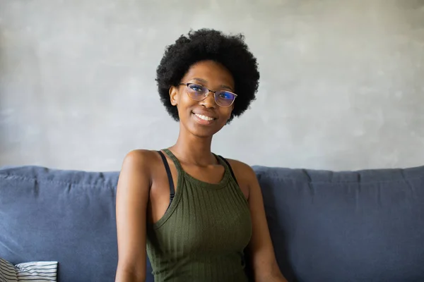 Happy african American young woman looking in camera sit relax on cozy couch happy to move to new apartment. Smiling black girl rest on comfortable sofa in living room dreaming