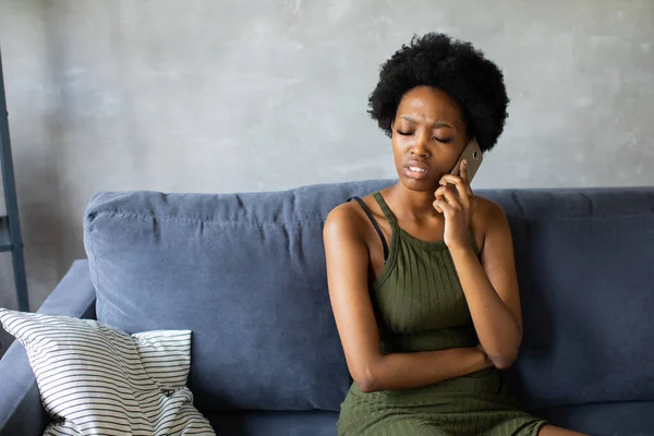 Unhappy African American woman talking on the phone, angry girl talking on the phone, discharged or broken mobile device, problem with phone, sitting on sofa