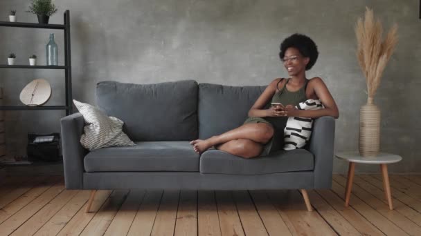 Feliz afroamericana joven mujer sentarse relajarse en acogedor sofá y feliz con el mensaje en el teléfono — Vídeos de Stock