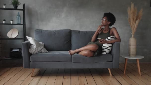 幸せなアフリカ系アメリカ人の若い女性は居心地の良いソファでリラックスし、電話でのメッセージに満足 — ストック動画