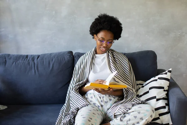 Happy African American young woman sit relax on cozy couch and reads a book. Happy to move to new apartment.