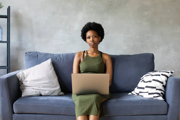 Happy african American young woman sit relax on cozy couch and working on a laptop. Happy to move to new apartment