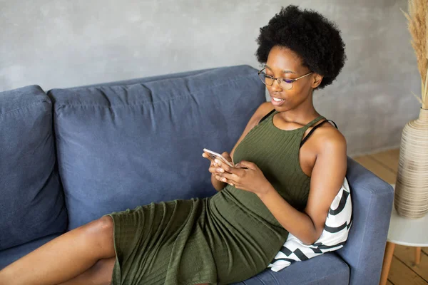 Happy african American young woman sit relax on cozy couch and writing on the phone. Happy to move to new apartment.