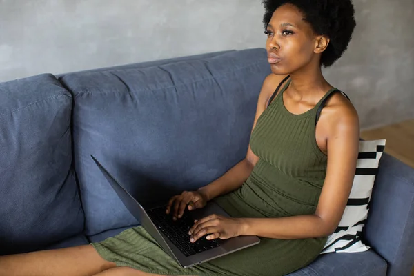 Happy african American young woman sit relax on cozy couch and working on a laptop. Happy to move to new apartment