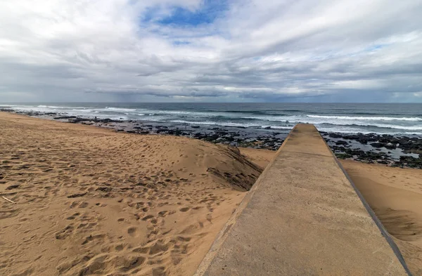 Concrete drainage pipeline extending onto sandy beach against rocky shoreline and blue cloudy coastal landscape at Garvies beach on the Bluff,  Durban, South Africa