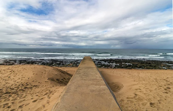 Concrete drainage pipeline extending onto sandy beach against rocky shoreline and blue cloudy coastal landscape at Garvies beach on the Bluff,  Durban, South Africa
