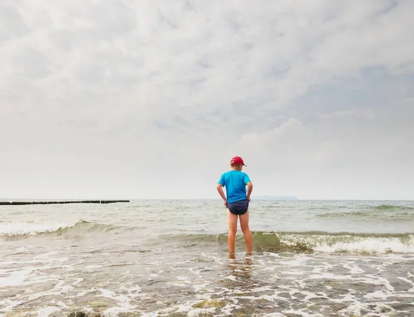 Boy in blue black sporty clothes is going into cold  water of sea. Windy day with foamy wavy sea. Blond hair  kid on stony beach.