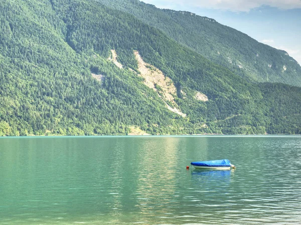 Boat on  green blue water level of Alps lake. White stony shore of a mountain lake. Reflection of forest mountains in water level.