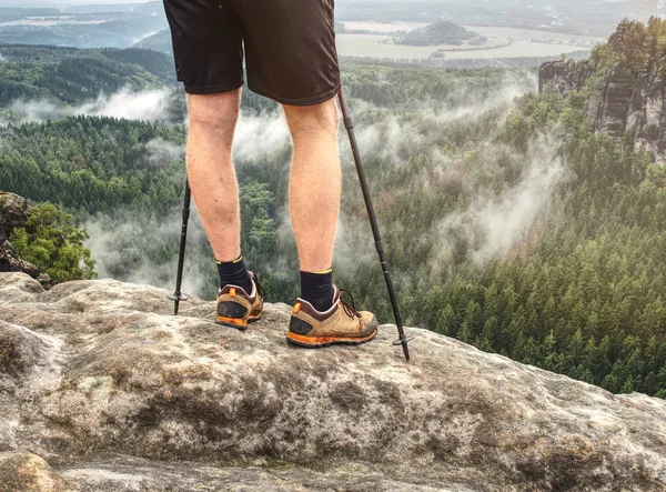 Nordic walking sticks and naked legs in sport shorts. Body  of a mountain hiker with hiking boots on a rock.