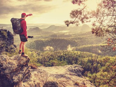 Ağır sırt çantası ve tripod elinde ile uzun boylu fotoğrafçı ayağa kayalık bakış ve derin puslu Vadisi feryat aşağı izlerken