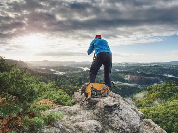 Photographer in blue sweatshirt working with mirror camera and tripod on peak of rock. Dreamy foggy landscape.