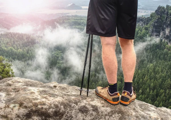 Nordic walking sticks and naked legs in sport shorts. Body  of a mountain hiker with hiking boots on a rock.