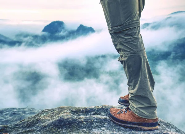 Hiker legs in light outdoor trousers and comfortable leather  trekking boots on rocky peak. Misty valley below peak in far blurry background. Hiking in nature.