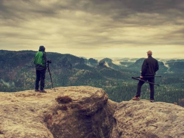 Fotoğrafçı fotoğraf makinesi tripod üzerinde kamera görünümünü kontrol edin. Adam uçurumun üzerinde kalmak ve konuşma arkadaşları fotoğraf çeker. Sonbahar güzel misty manzara puslu güneş doğarken ufuk. Dağlar erkeklerde