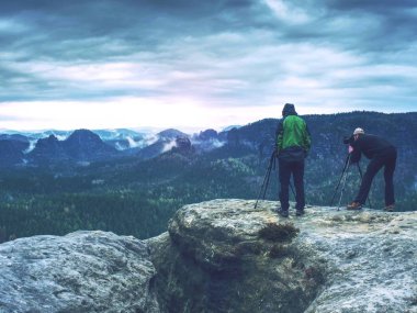 Fotoğrafçı fotoğraf makinesi tripod üzerinde kamera görünümünü kontrol edin. Adam uçurumun üzerinde kalmak ve konuşma arkadaşları fotoğraf çeker. Sonbahar güzel misty manzara puslu güneş doğarken ufuk. Dağlar erkeklerde