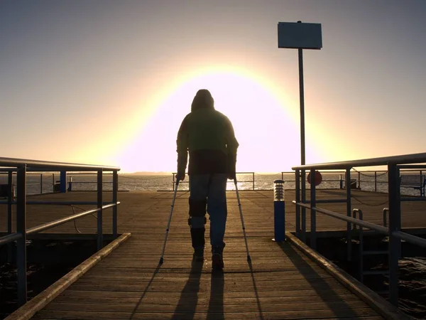 Hard walk of hurt man with crutches on port pier. Marwelous sunbreak at winter sea.