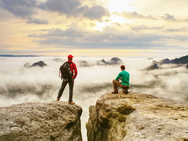 Two friends, hiker thinking and photo enthusiast takes photos of fall landscape. Cold and  mist weather.  Dreamy fogy landscape blue misty sunrise in a beautiful valley below