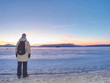 Sıcak uzun ceketli kadın donmuş göl veya nehir de kumlu plajda kalmak. Tepelik ufukta gün tatili ile kış soğuk sabah.