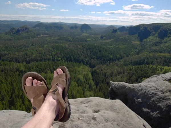 Male hairy legs with sandals rest on rocky summit above valley