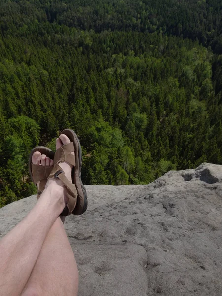 Male hairy legs with sandals rest on rocky summit above valley