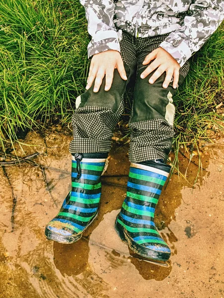 Criança usando botas de chuva, senta-se na poça enlameada — Fotografia de Stock