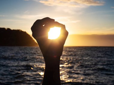 Gölge şeklindeki siluet, kumsalda güneş ışığı olan eller tarafından yapılıyor. Denizde gün batımı. Güneşe dokun. Yuvarlak çerçeveli parmaklar