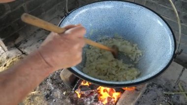 Bahçe mutfağında yemek pişiren, saksıya soğan ekleyen ve tahta kaşıkla dönen yaşlı bir adam. Yavaş çekim.