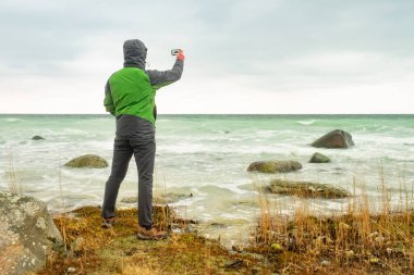 Yeşil yağmurluk giyen yürüyüşçü sahilde akıllı telefon kamerasıyla fotoğraf çekiyor..