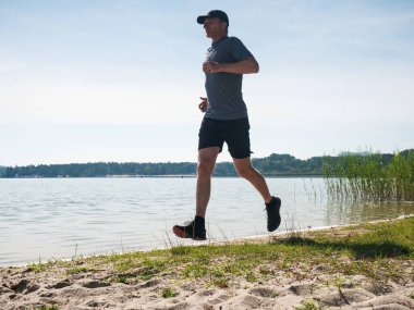 Beyzbol şapkalı ve kırmızı mavi spor kıyafetli uzun boylu adam plajda koşuyor ve egzersiz yapıyor. Dağ Gölü sahilinde aktif spor adamı. 