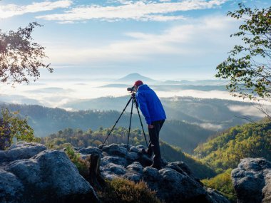 Görüntü yönetmenindeki fotoğraf çerçevesi sahnesi. Adam sisli dağların manzarasını çekmek için tripodun üzerinde kamera kullanıyor. Tatil konsepti seyahatinde dinlenme zamanı