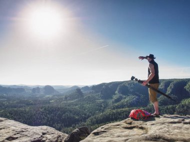 Fotoğrafçı, sol elinde kamerayla tripodu tutarken gözleri gölgeliyor. Fotoğrafçı kanyonun yukarısındaki kayalıklarda geziyor.