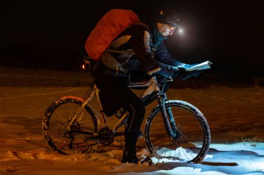 Night bike rider check the track itinerary in the light of a headlight. Lost path in snowdrift, snowflakes melting on dark off road tyre. Extreme bike orienteering race. clipart