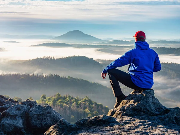 Man on the peak of rock. Man sit with hand on knee with beautiful sunrise landscape.  Hiker is watching into colorful mist and fog in forest valley.