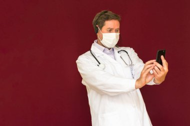 Stetoskop ve tıbbi maskeli beyaz bir doktor hasta ile çevrimiçi kırmızı stüdyo geçmişi hakkında konuşuyor.