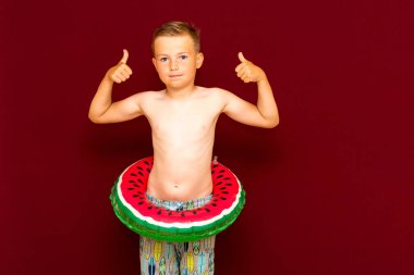 Yaz konsepti, mutlu beyaz çocuk öğrenci portresi çıplak mayo başparmağını kaldırıyor, karpuz şişirilebilir yüzük tutuyor, yaz tatilinde eğleniyor, stüdyoda kırmızı arka plan çekiliyor..